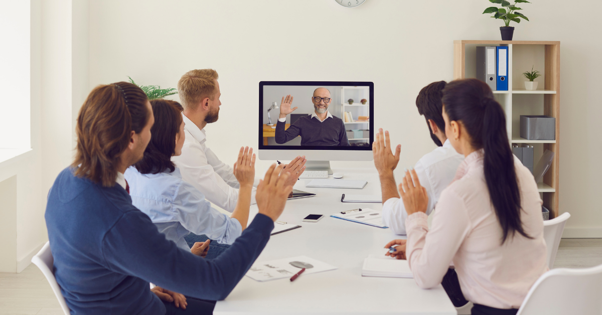 4 employees in office waving at man on laptop working virtually hybrid