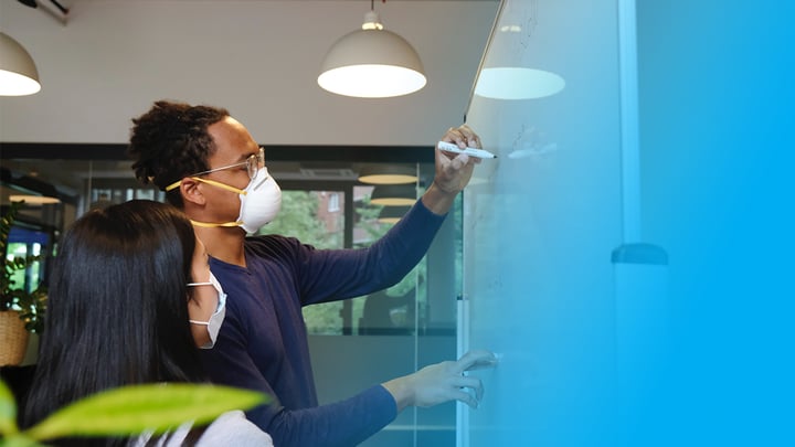 Employees wearing protective masks collaborate on a whiteboard in a post-COVID office environment.